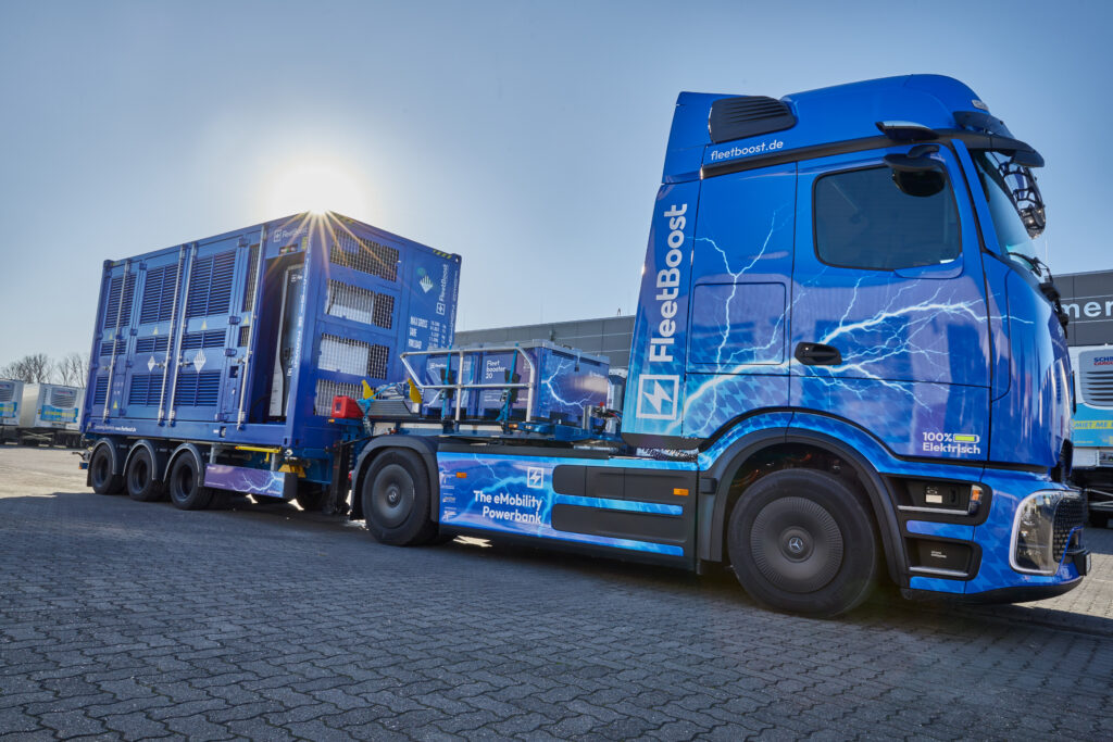 der Fleetbooster 20 wird von einem e-LKW gezogen. Der e-LKW it blau und auf ihm sind hellblau Blitze abgebildet. Das ganze ist aus der Froschperspektive fotografiert.